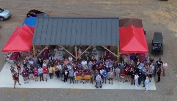 A large group of people seen from above stand before a picnic shelter and red-topped event tents.