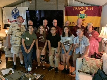 A group of young adults stand in foreground with a line of older adults behind them. All stand before a Norwich University flag to right, and US service flag to their left.