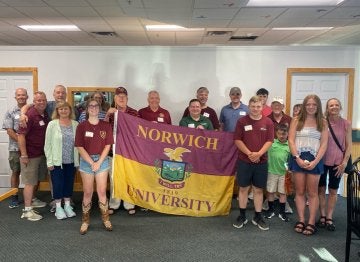 A group of young and older adults, with those in the center holding a maroon & gold Norwich University flag.