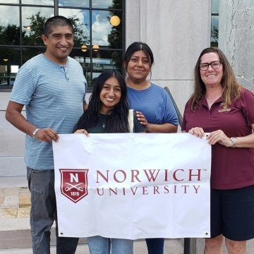 A small group of individuals stand and hold a white flag with Norwich University and school's shield "N" logo.