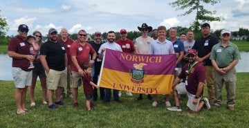A group of young and older adults stand outside, with those in the center holding a maroon & gold Norwich University flag.