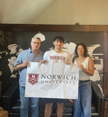 A group of three individuals holding a white with maroon lettering Norwich University flag.