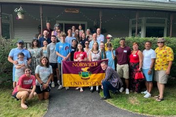 A group of young and older adults, with those in the center holding a Norwich University flag.