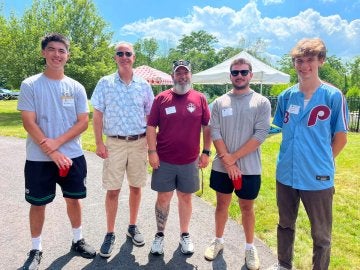 Five individuals stand outside on a mostly sunny day.