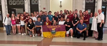 A sizeable crowd of individuals stand in three-to-four rows, with center individuals holding a maroon and gold Norwich flag.