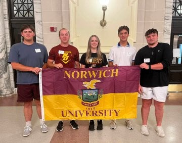 Five young adults stand indoors holding a Norwich University maroon & gold flag.