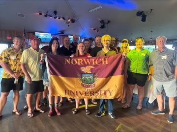 A group of young and older adults, with those in the center holding a maroon & gold Norwich University flag.