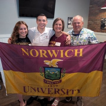 A group of four individuals holding a maroon & gold Norwich University flag.