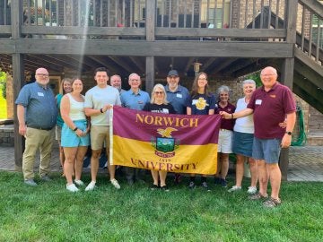 A group of young and older adults, with those in the center holding a Norwich University maroon & gold flag.