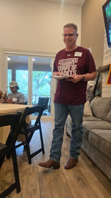 An individual with glasses wearing a maroon t-shirt holds a folder, standing between a table and a couch.