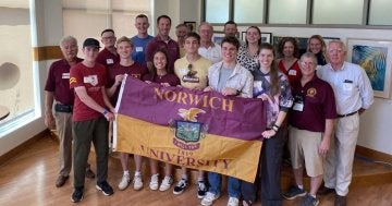 A group of young and older adults, with those in the center holding a maroon & gold Norwich University flag.