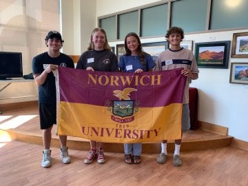 Four young adults hold a maroon & gold Norwich flag.