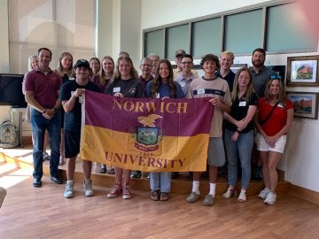 A group of young and older adults, with those in the center holding a Norwich University flag.